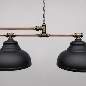 May include: A dual-pendant light fixture featuring two matte black dome shades suspended from a horizontal copper pipe frame. The structure is connected by dark bronze-toned metal pipe fittings and hangs from a central metal chain.
