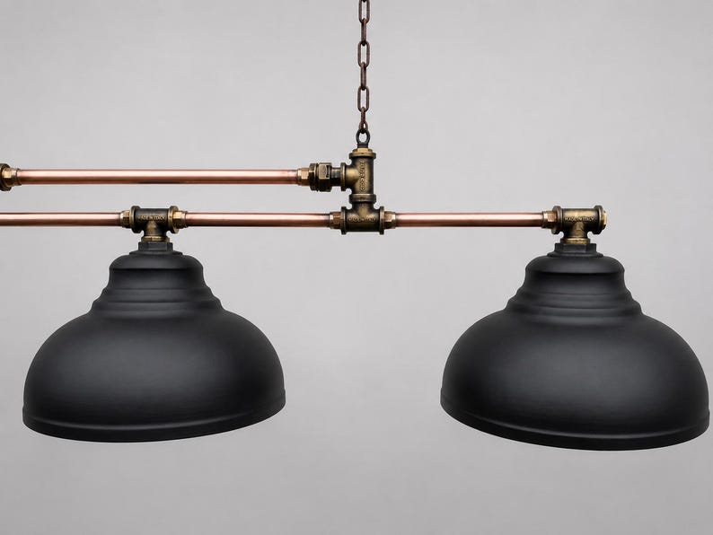 May include: A dual-pendant light fixture featuring two matte black dome shades suspended from a horizontal copper pipe frame. The structure is connected by dark bronze-toned metal pipe fittings and hangs from a central metal chain.