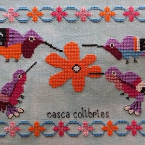 Peut inclure: Une broderie au point de croix représentant quatre colibris colorés autour d'une fleur orange. Les oiseaux sont dans des tons de violet, orange et rouge, avec des becs noirs. Le texte "nasca colibries" est en dessous, avec une bordure florale.
