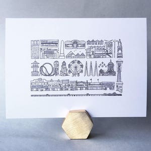 May include: Black and white line drawing print of the Seattle skyline, featuring iconic landmarks like the Space Needle, the Ferris wheel, and the pier. The print is on white paper and has a simple, minimalist style.