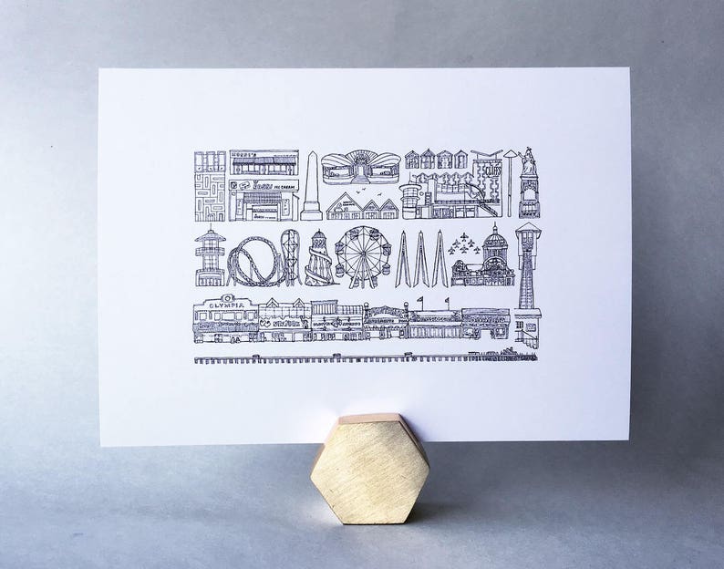May include: Black and white line drawing print of the Seattle skyline, featuring iconic landmarks like the Space Needle, the Ferris wheel, and the pier. The print is on white paper and has a simple, minimalist style.