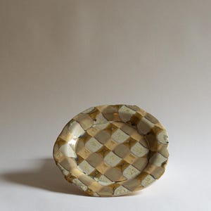 Może przedstawiać: Ręcznie robiony ceramiczny talerz z unikalnym wzorem w szachownicę w odcieniach beżu, jasnobrązowego i kremowego. Talerz ma nieregularną, lekko falistą krawędź, co dodaje mu uroku rzemieślniczego. Idealny do serwowania przystawek lub jako element dekoracyjny.