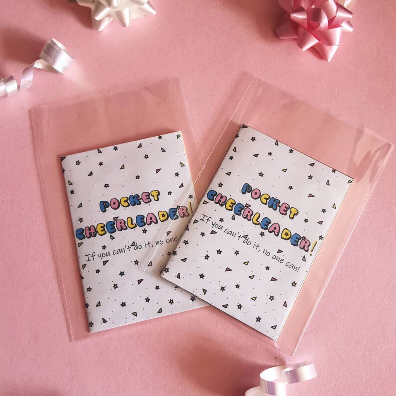 May include: Two small white pocket-sized notebooks with colorful text that reads "Pocket Cheerleader!" and "If you can't do it, no one can!" The notebooks are in clear plastic bags on a pink background.