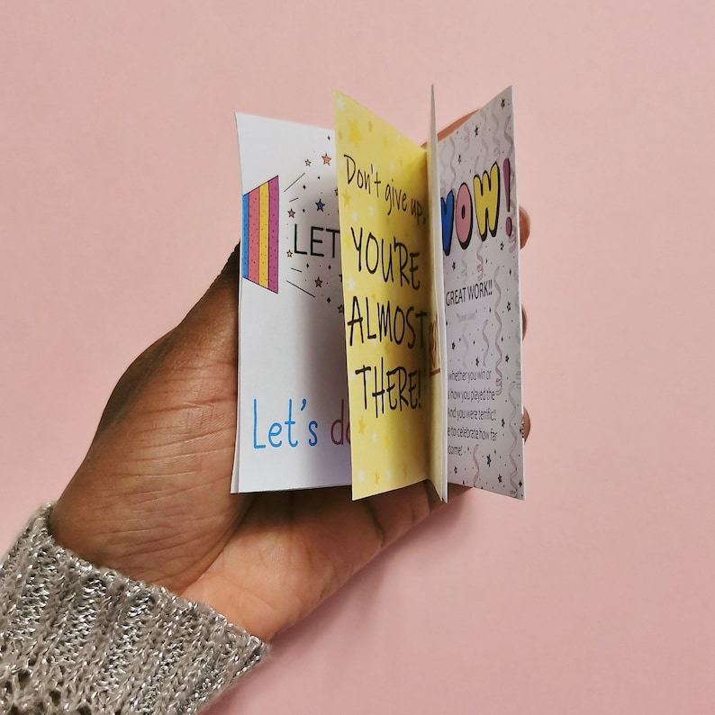 May include: A hand holding a set of three colorful motivational cards with encouraging messages. The cards feature bright colors, stars, and handwritten text. The text on the cards reads "Don't give up, you're almost there!" "Let's do great work!" and "Wow!"