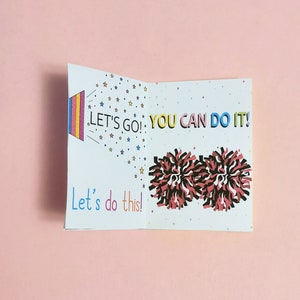 May include: A white card with a colorful design and the text "Let's go! You can do it! Let's do this!"  The card features a rainbow-colored megaphone with stars and two red and black pom-poms.