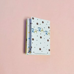 May include: A small, white booklet with a colorful star pattern on the cover. The text "RA-RA-RA" is written in blue and yellow letters on the front cover. The back cover has the text "honeybliss publishing.etsy.com".