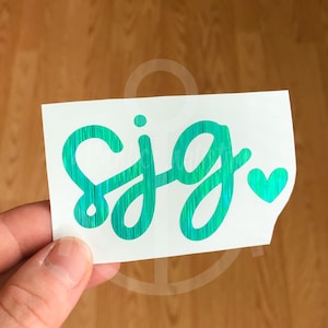 May include: A white sticker with the name "SJG" written in a turquoise holographic font with a small heart at the end.