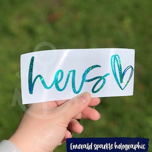 May include: A teal green holographic vinyl decal with the word "hers" and a heart. The decal is on a white background.