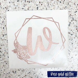 K&ouml;nnte beinhalten: Aufkleber aus ros&eacute;goldfarbenem Glitzer mit dem Buchstaben W in einem geometrischen Rahmen mit floralen Akzenten.