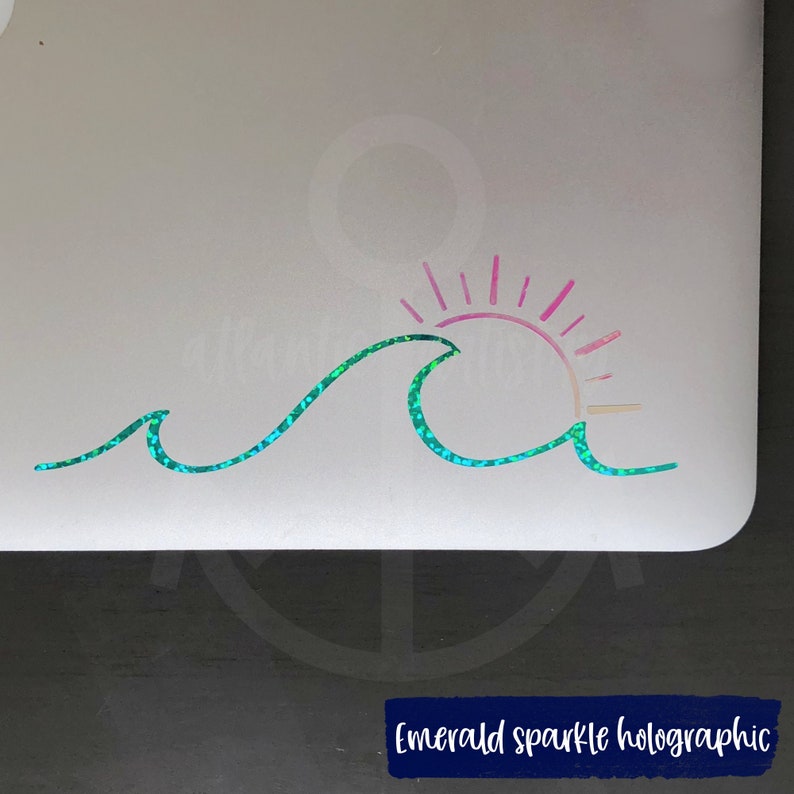 May include: A holographic decal featuring an emerald green wave and a pink and yellow sun. The design is applied to a light grey surface. The text "Emerald sparkle holographic" is displayed at the bottom. The wave and sun design is a simple, stylized illustration.