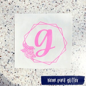 K&ouml;nnte beinhalten: Ein pinkfarbener Glitzer-Vinyl-Aufkleber mit einem geometrischen Rahmen und einem floralen Design. Der Buchstabe "G" befindet sich in der Mitte des Rahmens.
