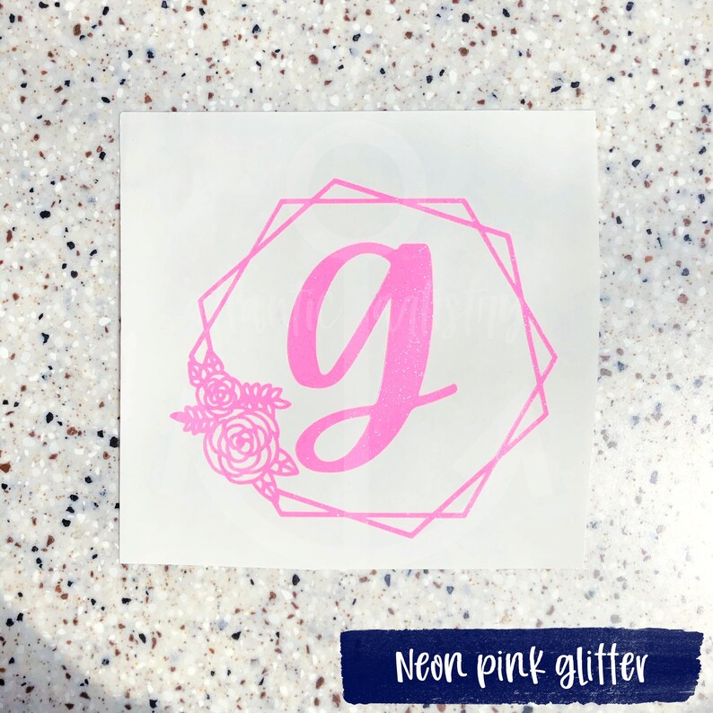 K&ouml;nnte beinhalten: Ein pinkfarbener Glitzer-Vinyl-Aufkleber mit einem geometrischen Rahmen und einem floralen Design. Der Buchstabe "G" befindet sich in der Mitte des Rahmens.