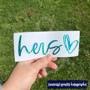 May include: A white vinyl sticker with the word "hers" written in a cursive font and a heart next to it. The sticker is made with emerald sparkle holographic vinyl.