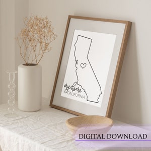 May include: A framed print featuring a black outline of the California state map with a heart symbol and the words "my home California" in a handwritten font. The frame is light brown, and the print is on a white background.