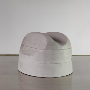 Puede incluir: Una escultura de hormigón redondeada, de color gris claro, con una parte superior curvada y una ranura. La escultura tiene líneas horizontales y un acabado mate y liso. El fondo es una pared blanca lisa.