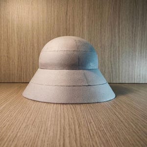 Puede incluir: Un expositor de sombreros de espuma gris claro en forma de cúpula con un ala ancha y acampanada. La estructura presenta dos costuras horizontales distintas que rodean la copa y el ala, creando un aspecto escalonado con textura mate.