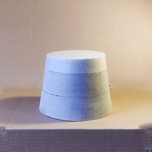 Puede incluir: Objeto cilíndrico azul claro hecho a mano, con una parte superior plana y una base ligeramente más ancha. La superficie es texturizada y parece estar hecha de hormigón o material similar. El diseño incluye líneas horizontales.