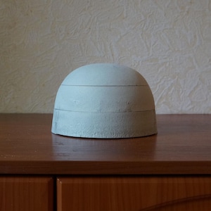 Puede incluir: Un objeto gris claro en forma de cúpula con tres líneas horizontales. El objeto está sobre una superficie de madera marrón oscuro. El fondo es una pared texturizada de color beige claro.