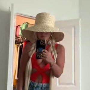 May include: A woman wearing a red one-piece swimsuit with cutouts, blue denim shorts, and a wide-brimmed straw hat. She is taking a selfie with a phone case that has a blue and gold design.