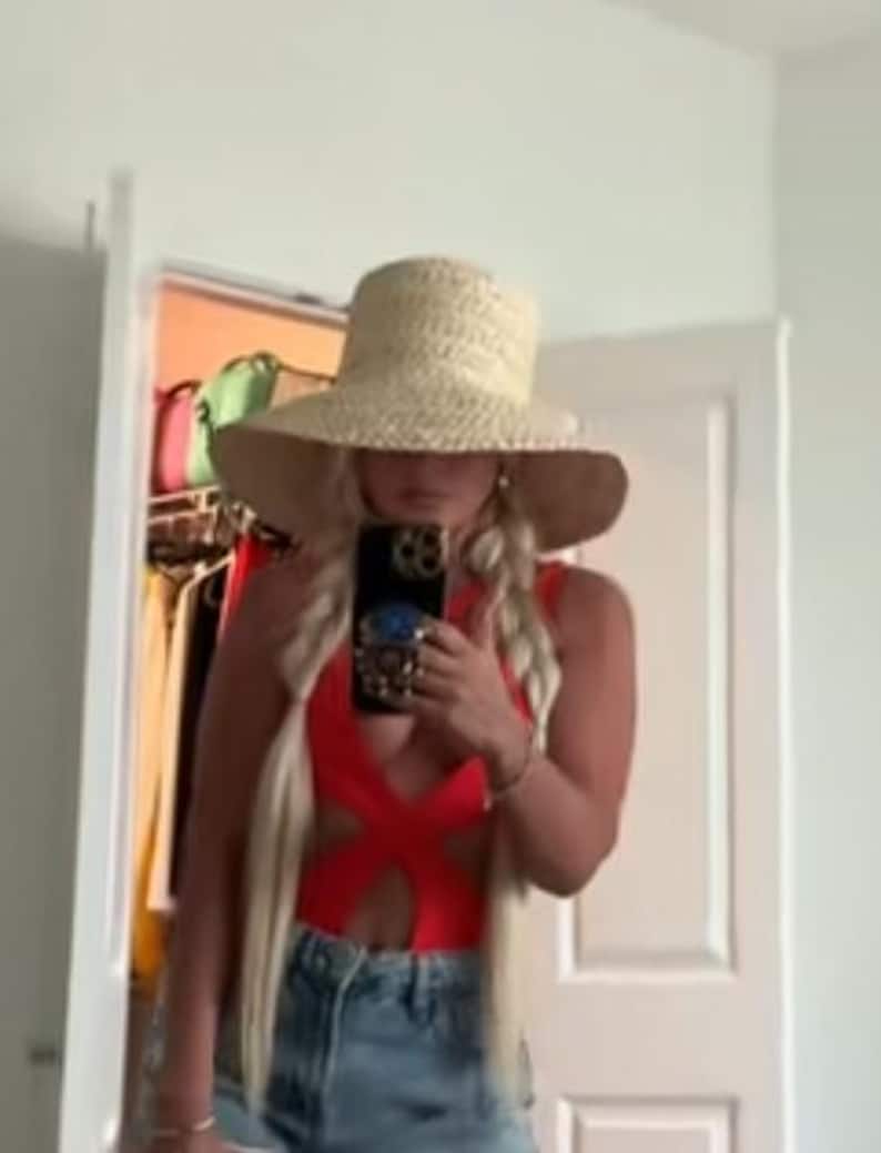 May include: A woman wearing a red one-piece swimsuit with cutouts, blue denim shorts, and a wide-brimmed straw hat. She is taking a selfie with a phone case that has a blue and gold design.