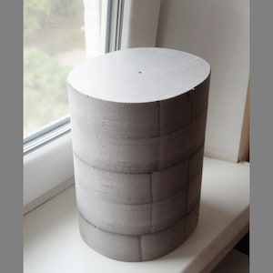 Puede incluir: Un cilindro de espuma gris con un patrón de cuadrícula en la superficie. El cilindro tiene aproximadamente 25 cm de alto y 15 cm de diámetro.