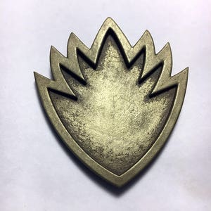 May include: A gold-colored metal pin in the shape of a stylized flame. The pin has a textured surface and a slightly worn look.
