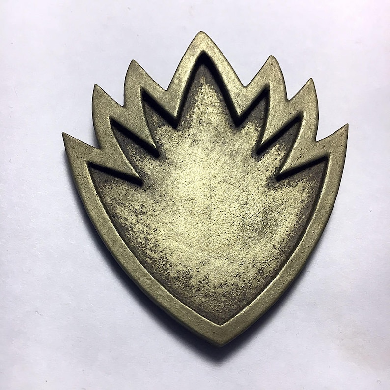 May include: A gold-colored metal pin in the shape of a stylized flame. The pin has a textured surface and a slightly worn look.
