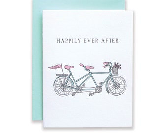 May include: A white card with the text "Happily Ever After" in black lettering. A hand-drawn illustration of a blue tandem bicycle with pink seats and a pink flag that says "Just Married" is below the text.