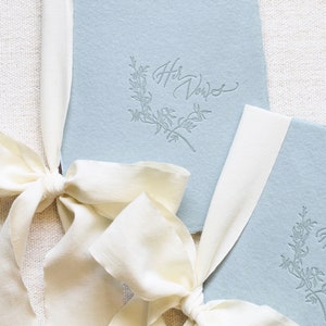 May include: Two light blue paper vow books with white ribbon bows. The top book has the text "Her Vows" in cursive script with a floral design.