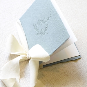 May include: A light blue book with the words "Our Vows" embossed on the cover. The book is tied with a white ribbon.