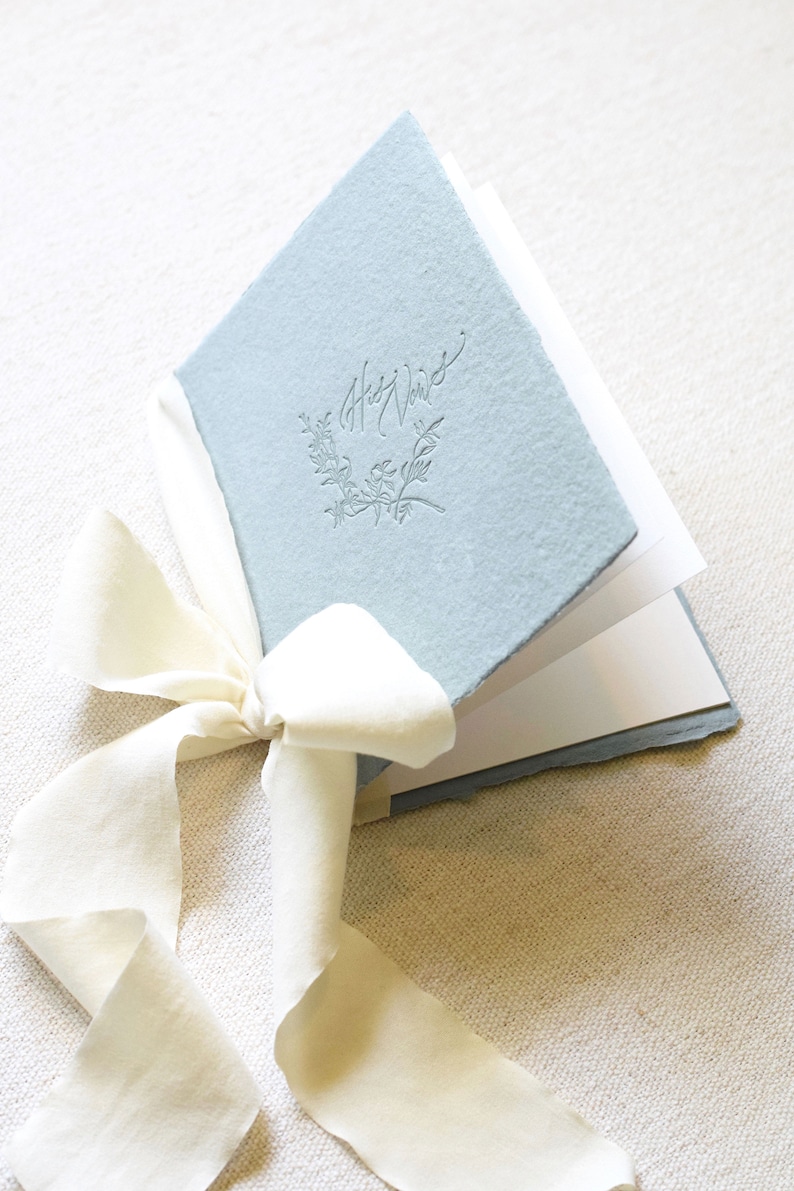 May include: A light blue book with the words "Our Vows" embossed on the cover. The book is tied with a white ribbon.