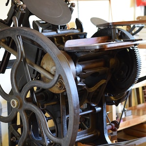 May include: A vintage printing press with a large wheel and a metal frame. The press has a wooden platform and a metal plate.