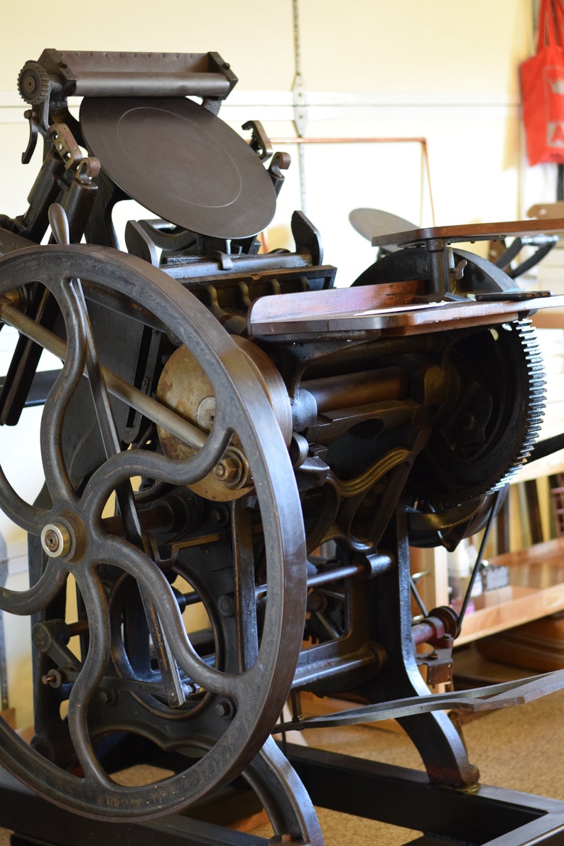 May include: A vintage printing press with a large wheel and a metal frame. The press has a wooden platform and a metal plate.