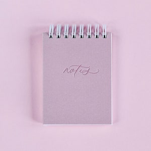 May include: A pink spiral bound notebook with the word "notes" written in cursive on the front cover.
