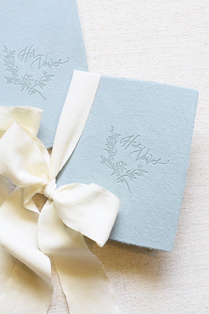 May include: Two light blue paper covered books with a white ribbon tied around them. The books have the text "Her Vows" and "His Vows" embossed on the covers.