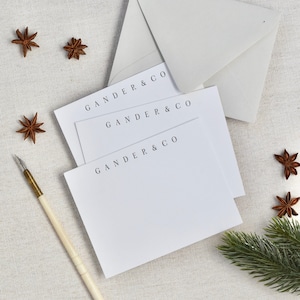 May include: A stack of white stationery cards with the text "GANDER & CO" printed on each card. The cards are on a beige linen surface with a sprig of evergreen and star anise.