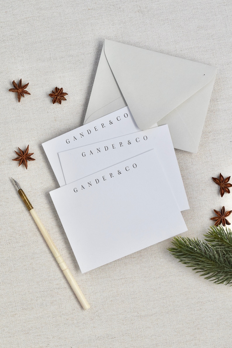 May include: A stack of white stationery cards with the text "GANDER & CO" printed on each card. The cards are on a beige linen surface with a sprig of evergreen and star anise.