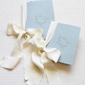 May include: Two light blue vow books with white ribbon bows. The books are tied together with the ribbons. The books have the text "Her Vows" embossed on the front.