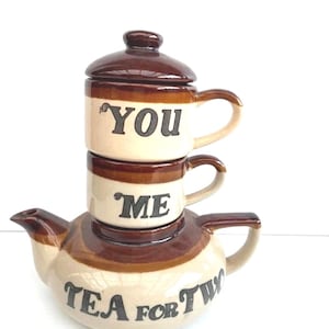 May include: A ceramic tea for two set featuring a teapot base and two stacked cups with lids. The cream-coloured pieces have brown glazed rims and bases. The top cup says YOU, the middle cup says ME, and the teapot base says TEA FOR TWO in black.