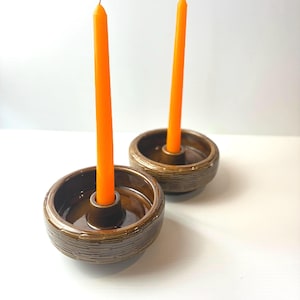 May include: Two round, brown ceramic candle holders with a textured, basket-weave pattern on the exterior. Each holder features a central spike for securing a single, slender, bright orange taper candle. The glaze is smooth and glossy.