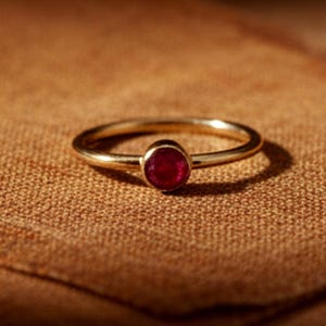 Op de afbeelding: Een gouden ring met een kleine, ronde, rode edelsteen. De ring ligt op een bruin stoffen oppervlak, wat een contrast creëert tussen het glanzende metaal en de textuur van de achtergrond. De ringband is dun en delicaat.
