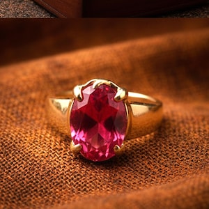 May include: A gold ring with a large, oval-cut, ruby-red gemstone. The ring's band is a polished gold, and the gemstone is set with four prongs. The ring is displayed on a brown, textured surface.