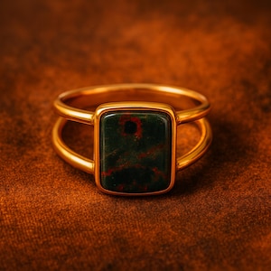 Peut inclure: Une bague dorée avec une pierre rectangulaire vert foncé avec des inclusions rouges. La bague a un design à double bande, la pierre étant sertie dans une lunette dorée. La bague est présentée sur une surface marron texturée.