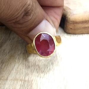 May include: A gold ring featuring a large oval ruby gemstone. The ruby is a deep red colour and has a faceted cut.