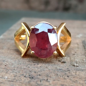 Puede incluir: Un anillo de oro con una gran piedra preciosa de rubí de forma ovalada. El anillo tiene un diseño único con dos bandas de oro curvadas que se unen en la parte superior e inferior del rubí.