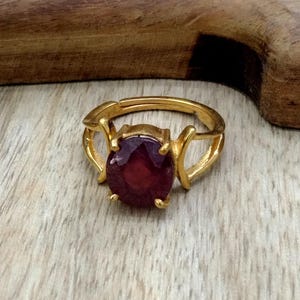 May include: A gold ring with a large, round, dark red gemstone set in a prong setting. The ring has a delicate, openwork design.