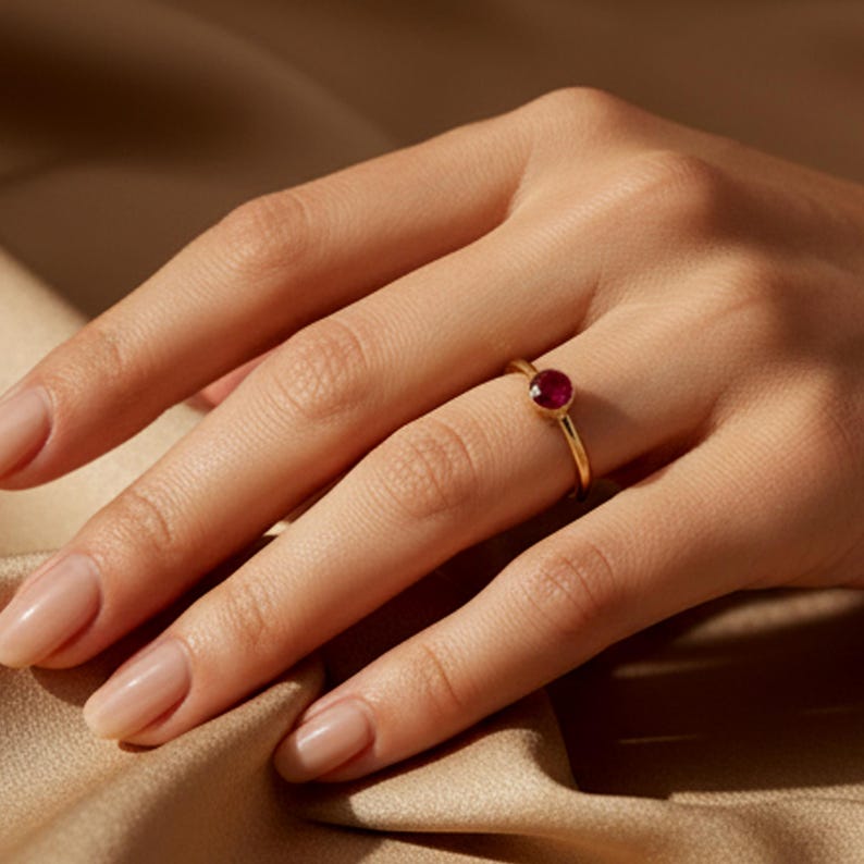 May include: A gold ring featuring a small, round, red gemstone. The ring is worn on a finger. The background is a soft, neutral tone, accentuating the ring's design and colour. The ring's gemstone is a deep red.