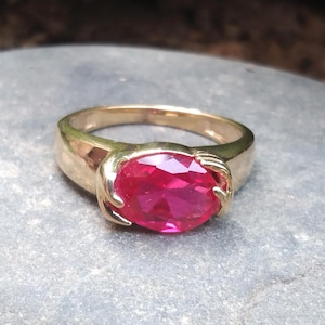 Peut inclure: Une bague en or avec une grande pierre précieuse ovale de couleur rose vif sertie au centre. La bande de la bague est conçue pour ressembler à deux mains tenant la pierre précieuse.
