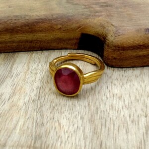 May include: A gold ring featuring a large, round, red gemstone set in the centre. The ring has a simple, elegant design.