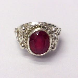 May include: A silver ring with an oval, faceted ruby gemstone. The ring has a decorative design with rope and swirl details around the gemstone. The ruby is a deep red colour, and the ring is set against a white background.